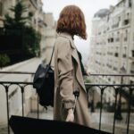 Woman in trench coat walking through a Parisian street with an umbrella, showcasing city life and travel.