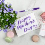 Flat lay of Mother's Day card with purple flowers, gift, macarons, and a pen indoors.