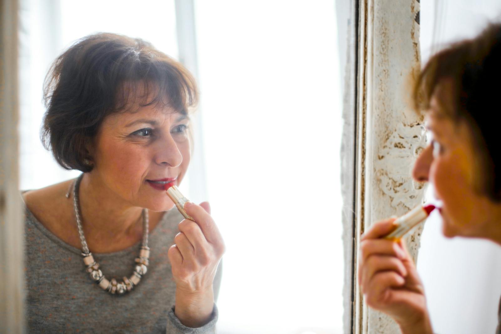 Elegant senior woman applying lipstick while looking in the mirror indoors.